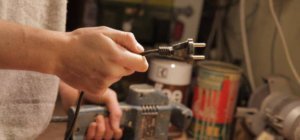 Ein Mann hält in seiner rechten hand einen Stecker. Er hält ihn vor sich. er trägt einen grauen Pullover. Mit seiner linken Hand hält er eine Schleifmaschine fest, die auf einem Tisch zu stehen scheint. Im Hintergrund ist ein wenig Chaos.