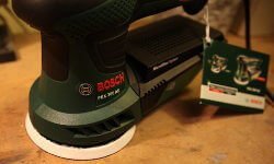 Zu sehen ist das Gehäuse des Exzenterschleifers PEX 300 AE. Es besteht aus Plastik. Das Bosch Logo ist in silber und die Bosch Schrift in rot. Die Schleifplatte in weiß gehalten. Außerdem sieht man die Staubkassette und eine Beschreibung die an dem Griff hängt.