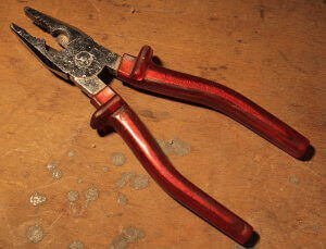 Ein geöffneter Seitenschneider mit rotem Griff liegt auf einem älteren Holz Untergrund.Der Griff ist dunkelrot. Die Marke ist nicht erkennbar.
