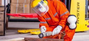 Ein Handwerker schleift ein Stück Metall. Er ist in voller Sicherheitsmontur gekleidet. Er trägt einen orangenen Ganzkörperanzug, Knieschoner, einen Sichtschutz, Ohrschutz und einen Mundschutz. Er schleift mit einem Winkelschleifer. Alle Farben die er trägt sind hell.