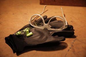 Eine Schutzbrille, Handschuhe und zwei Ohr stopfen liegen beieinander. Sie liegen auf einem Hölzernen Untergrund.