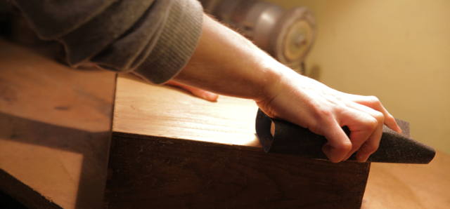 Die Hand eines jungen Mannes greift um einen Schleifblock, der umwickelt ist mit schwarzen, grobem Schleifpapier. Er schleift gerade eine Holzkiste. Dabei wird die Oberfläche abgeschliffen.