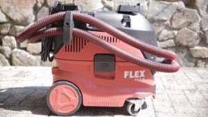 Von der Seite ist der Industriestaubsauger VCE 33 MAC von der Firma Flex zu sehen. Er ist in den Farben Rot, Schwarz und Weiß gehalten. Er steht vor eine Natur Steinwand und auf einem gepflasterten Boden. Das Hinterrad ist um einiges größer als das Vorderrad. Der Schlauch ist mehrfach um den Industriestaubsauger gewickelt.